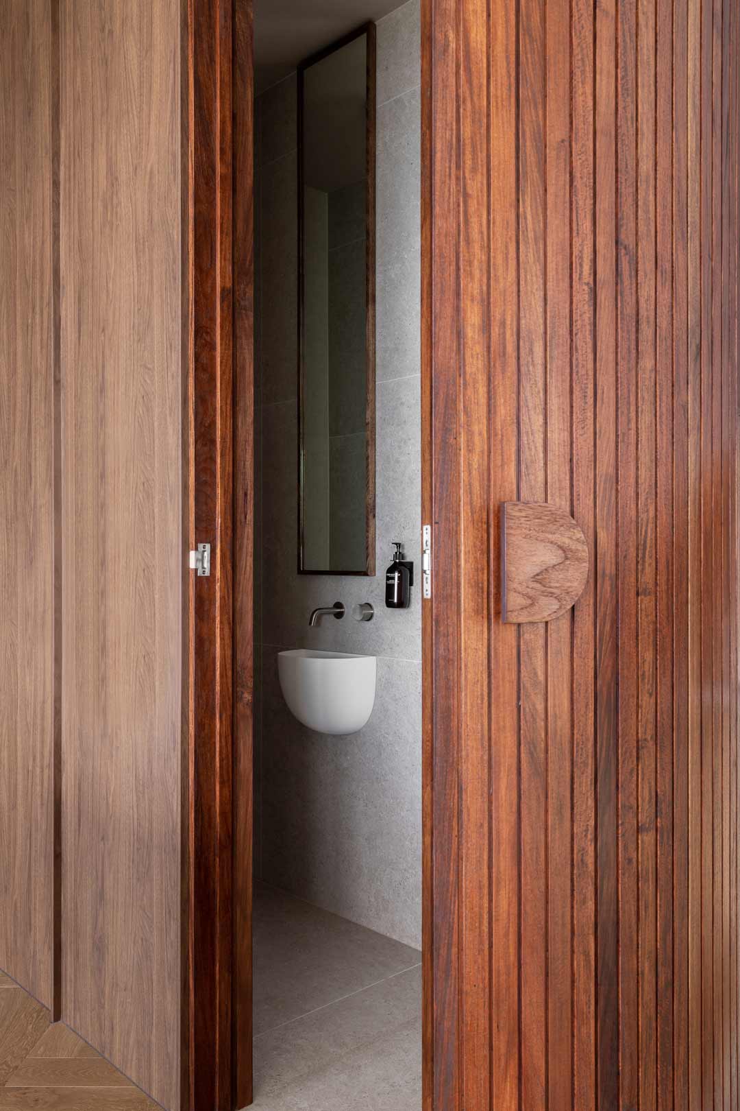 A-hidden-powder-room-through-walnut-timber-fluted-doors-with-a-small-wall-hung-basin-and-wall-mounted-tapware